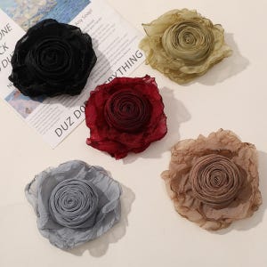 May include: Five fabric flower embellishments in black, olive green, burgundy, light blue, and tan. Each flower has a ruffled, layered design, resembling a rose. The flowers are arranged on a light-coloured surface.
