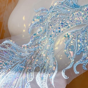Puede incluir: Primer plano de una tela bordada en azul claro con lentejuelas. El diseño presenta patrones fluidos en forma de pluma con intrincados abalorios y lentejuelas iridiscentes. La tela está montada sobre un respaldo de malla blanca, creando una apariencia delicada y texturizada.