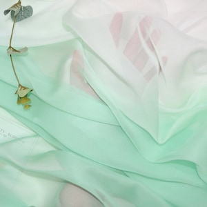 Puede incluir: Una tela transparente de color verde claro con un degradado blanco. La tela está drapeada y doblada, creando líneas suaves y fluidas.