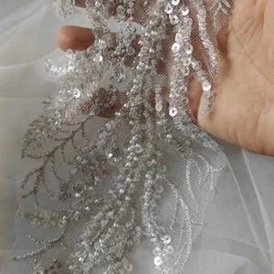 May include: Close-up of silver lace trim with intricate beaded and sequined floral design. The delicate trim features a net backing and is embellished with shimmering beads and sequins, creating a luxurious and elegant look. Ideal for embellishing garments or accessories.