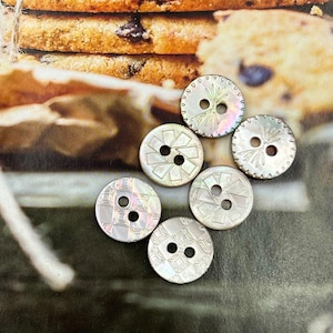 Może przedstawiać: Sześć okrągłych, iryzujących guzików z masy perłowej, każdy z dwoma otworami. Guziki mają różne wytrawione wzory, w tym geometryczne wzory i motywy kwiatowe. Guziki są rozrzucone na powierzchni z rozmytym tłem ciasteczek.