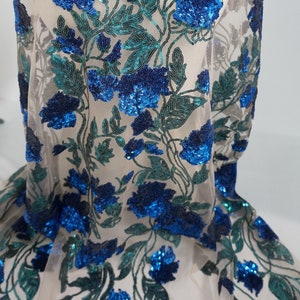 May include: Sheer fabric with a floral design. The flowers are a vibrant blue, created with sequins, and the leaves are a teal green. The fabric is draped over a form, showcasing the intricate embroidery and texture of the design.