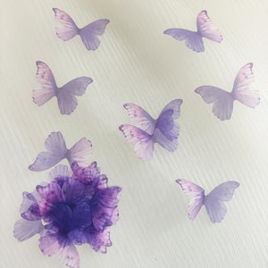 Könnte beinhalten: Eine Ansammlung von durchscheinenden hellvioletten Schmetterlingsausschnitten, die um ein Bündel geschichteter dunkelvioletter Stoffblütenblätter angeordnet sind. Die Schmetterlinge weisen zarte Flügelmuster auf und sind auf einer strukturierten cremeweißen Oberfläche verteilt.