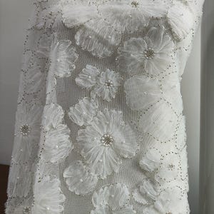 Puede incluir: Una tela blanca transparente con aplicaciones florales tridimensionales hechas de pétalos de organza en capas. El centro de cada flor está adornado con pequeñas perlas blancas y cuentas transparentes, con abalorios plateados.