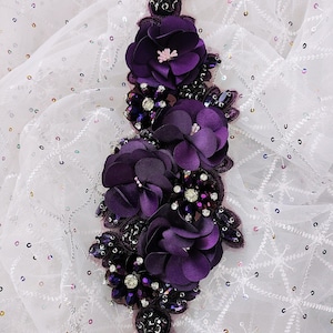 Puede incluir: Una aplicación decorativa vertical con flores de tela de color morado intenso, superpuestas con cuentas de cristal, lentejuelas y detalles metálicos. El arreglo floral está montado sobre una base de malla oscura con extremos puntiagudos.