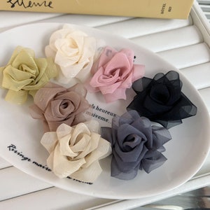 May include: Assortment of fabric flower embellishments in various colors, including beige, pink, brown, black, and gray. The flowers are arranged on a white plate.