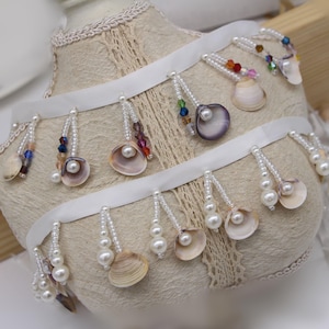 以下が含まれることがあります： 真珠と貝殻のアクセントが付いた白いサテンリボン飾り。トリムには、白い真珠と小さな貝殻の列があり、2列目はカラフルなビーズと大きな貝殻が並んでいます。