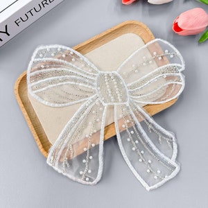 Può includere: Un fiocco bianco in tulle con decorazioni di perle. Il fiocco è su una superficie di legno.