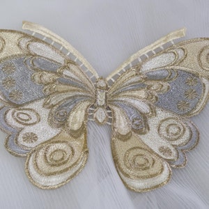 May include: A gold and silver embroidered butterfly applique with intricate details. The butterfly has a light brown background with gold and silver swirls and patterns.
