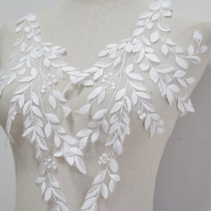 May include: White floral lace applique with a detailed leaf and berry pattern. This applique is crafted from a soft, textured material, perfect for enhancing bridal gowns, dresses, or craft projects. The design is symmetrical and elegant.