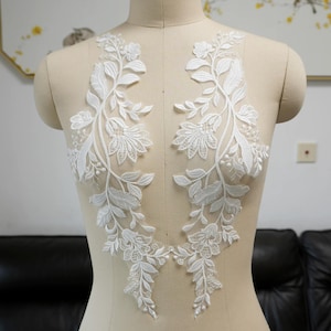 May include: A pair of white lace appliqués featuring intricate floral and leaf embroidery patterns. The symmetrical designs are crafted with fine thread detailing on a sheer mesh base, creating a delicate botanical motif for garment decoration.