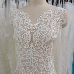 May include: White lace applique with detailed floral and scroll patterns. The applique has a V-neck shape and is decorated with sequins. This delicate piece is likely for bridal or formal wear.