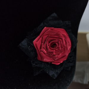 Puede incluir: Una rosa roja de satén, adornada con un cuadrado de encaje negro. La rosa es el centro, con el encaje como fondo. Este elemento decorativo es probablemente un accesorio, como un broche o un adorno.