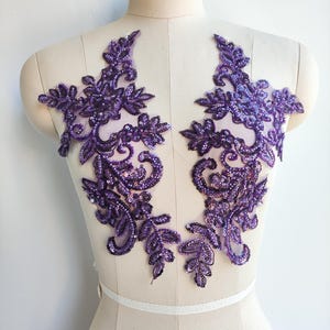 May include: Two symmetrical, purple sequined lace appliques. Each applique features a floral and leaf design with intricate detailing. The appliques are displayed on a mannequin, showcasing their potential use in garment embellishment or craft projects.