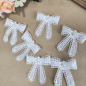 Puede incluir: Seis lazos de encaje blanco con un diseño calado delicado están dispuestos sobre una superficie neutra. Cada lazo presenta una pequeña perla redonda en el centro, añadiendo un toque de elegancia. Ideales para adornar manualidades o ropa.