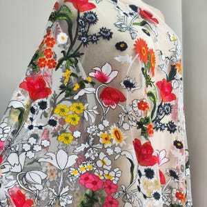Könnte beinhalten: Transparenter Stoff mit lebendiger Blumenstickerei. Das Design zeigt rote, gelbe und weiße Blumen mit grünen Blättern, wodurch ein farbenfrohes und detailliertes Muster entsteht. Der Stoff ist über einer Schaufensterpuppe drapiert, um die Textur und das Design hervorzuheben.