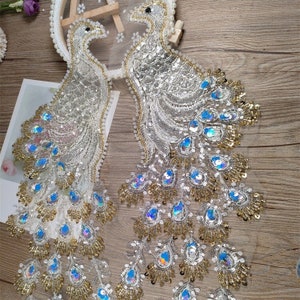 May include: Two white and gold peacock embroidery patches with iridescent blue sequins. The patches are on a white mesh fabric.