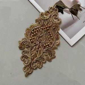 May include: Gold-colored embroidered applique with an ornate, symmetrical design. The applique features intricate patterns, small beads, and a central embellishment. The design is set against a neutral background, highlighting the detailed craftsmanship.