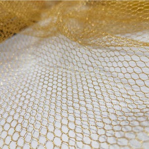 May include: Close-up of a gold hexagonal mesh fabric. The fabric has a delicate, net-like appearance with a subtle shimmer. The background is a soft, off-white colour, providing contrast to the gold mesh.
