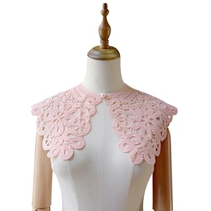 May include: A light pink lace collar with a delicate floral design. The collar is displayed on a mannequin and features a scalloped edge and a single pearl button closure at the front. This vintage-inspired accessory adds a touch of elegance.