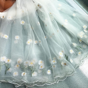May include: Delicate white sheer fabric adorned with embroidered daisy flowers. The fabric, with a scalloped edge, is draped over a pale blue surface. The daisies feature white petals, yellow centres, and green stems and leaves.