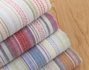 May include: A stack of four folded fabric pieces featuring vertical stripes in various colors including red, blue, green, yellow, and brown. Each piece displays a woven texture with repeating dashed lines and solid bands of color across the surface.