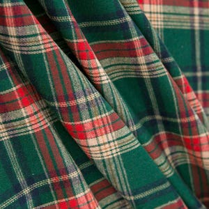 Puede incluir: Una tela con un patrón de cuadros escoceses clásico con líneas entrecruzadas en verde bosque profundo, rojo vibrante y crema. El material está drapeado en pliegues suaves, resaltando la textura tejida y el diseño de cuadrícula geométrica.