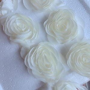 Può includere: Cinque fiori di tessuto bianco con bordi arricciati. I fiori sono disposti in fila su una superficie bianca.