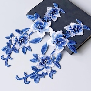 May include: Embroidered floral appliques in shades of blue and white. The appliques feature detailed flower and leaf designs, ideal for embellishing clothing or craft projects. The appliques are arranged on a dark grey surface.