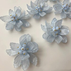 Peut inclure: Plusieurs ornements en forme de fleurs bleu clair à l'aspect translucide. Chaque fleur a de multiples pétales et un groupe de petites perles bleu clair avec des accents dorés au centre. Les fleurs sont dispersées sur une surface blanche.