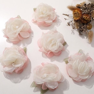 Puede incluir: Seis adornos florales artificiales con pétalos de tela translúcida superpuestos en blanco y rosa suave. Cada flor tiene un pequeño grupo de cuentas blancas en el centro y hojas de tela verde visibles en la base del arreglo floral.