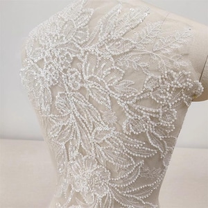 May include: White floral lace applique with intricate beadwork. The delicate design features a pattern of leaves and flowers, perfect for embellishing bridal wear or other garments. The lace is displayed on a mannequin.