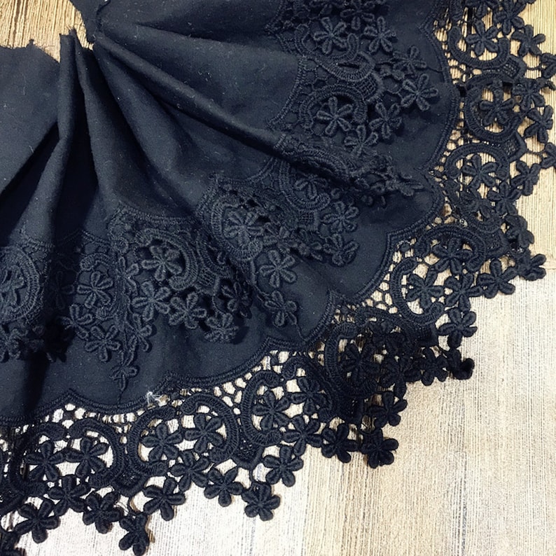 May include: Black lace trim with a scalloped edge and floral designs. The lace is folded in a fan shape and is on a wooden surface.