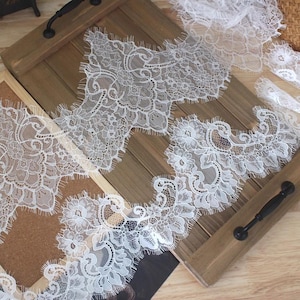 May include: White floral lace trim with scalloped edges. The delicate lace features intricate patterns of flowers and leaves. The trim is displayed on a wooden tray with black handles, showcasing its texture and design.