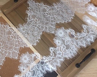 May include: White floral lace trim with scalloped edges. The delicate lace features intricate patterns of flowers and leaves. The trim is displayed on a wooden tray with black handles, showcasing its texture and design.