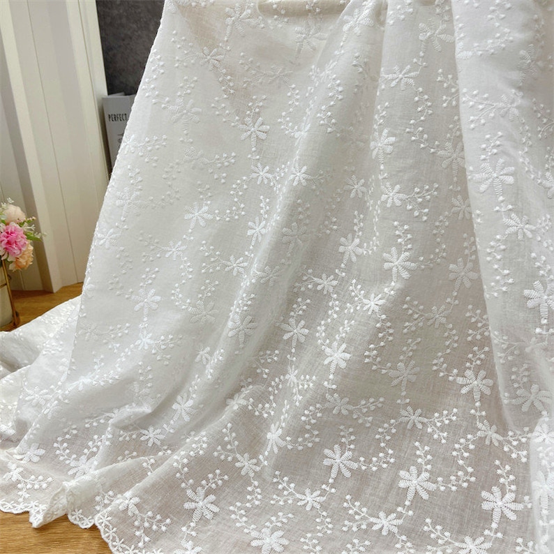 Puede incluir: Una tela blanca que presenta un intrincado patr&oacute;n floral bordado con delicadas enredaderas y peque&ntilde;as flores. El material es ligero y semitransparente, mostrando un dise&ntilde;o bot&aacute;nico repetitivo en toda la superficie del textil.