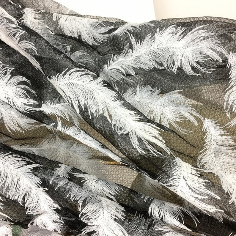 May include: Black sheer fabric with white embroidered feathers. The fabric is draped and folded, showing the texture of the mesh and the detail of the feathers.