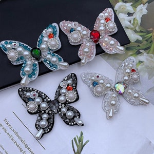 Pode incluir: Quatro enfeites em forma de borboleta com detalhes de pérolas e cristais. As borboletas são nas cores rosa, azul, preto e branco.