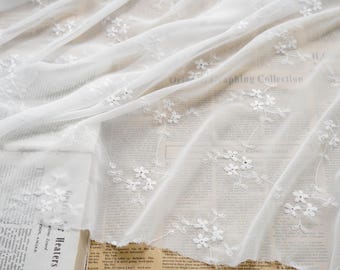 Puede incluir: Tela blanca bordada con motivos florales. El material transparente está drapeado, revelando un vistazo del texto impreso debajo. Delicadas flores y hojas blancas están bordadas en la tela, creando un patrón texturizado.