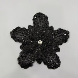 May include: A black sequined flower applique with a central rhinestone. The flower has five petals and is densely covered in small, shiny sequins. Ideal for embellishing clothing or accessories.