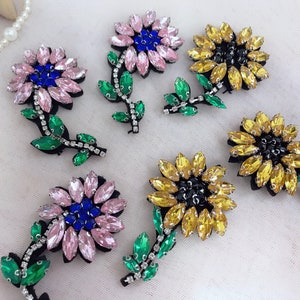 Puede incluir: Cinco adornos de flores de pedrería con hojas verdes. Las flores son rosas, amarillas y negras con pedrería azul en el centro.