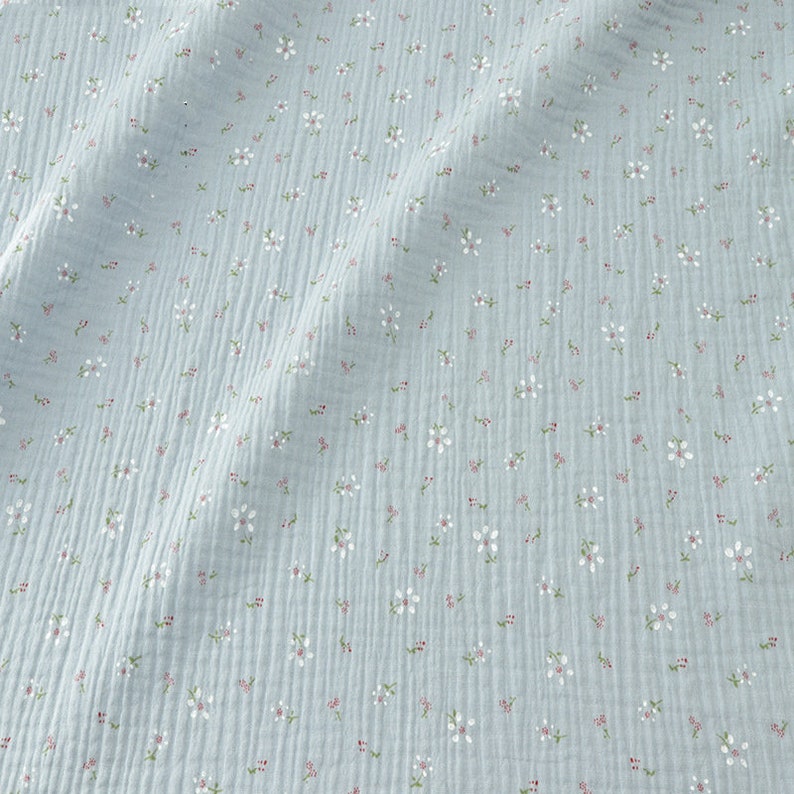 May include: Light blue fabric with a delicate floral pattern. The fabric features small white flowers with pink centers and green stems scattered across its surface. The fabric has a textured, crinkled appearance.