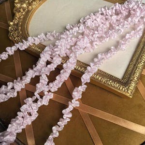 May include: Pale pink ruffled ribbon, ideal for crafting and embellishments, cascades across a gold-framed mirror. The ribbon's delicate texture and soft color create a vintage aesthetic. The background features a velvet surface and gold ribbon.