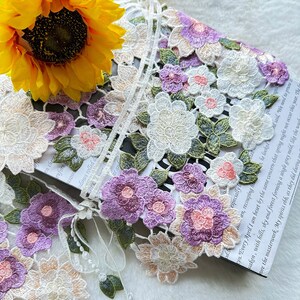 May include: White lace trim featuring embroidered flowers in shades of pink, purple, and green. The trim has a delicate floral pattern with a scalloped edge.