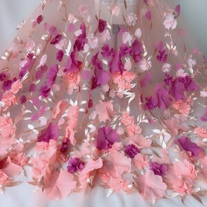 May include: Pink and purple 3D floral lace fabric with a delicate mesh background. The fabric features a gradient of colors, with pink flowers at the bottom and purple flowers at the top.