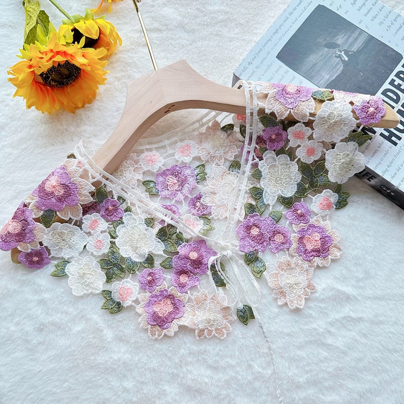 May include: White lace collar with embroidered flowers in shades of pink, purple, and green. The collar has a tie closure.