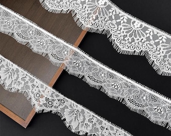 May include: Three strips of white lace trim featuring a detailed floral pattern with scalloped edges and delicate eyelash fringe. The lace is arranged in parallel lines, showcasing the intricate mesh texture and repeating botanical motifs.