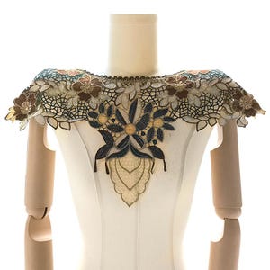 May include: A delicate lace collar featuring a floral design in shades of brown, blue, and white. The collar is crafted from fine, intricate lace fabric and showcases a central motif of a large flower with smaller flowers surrounding it.