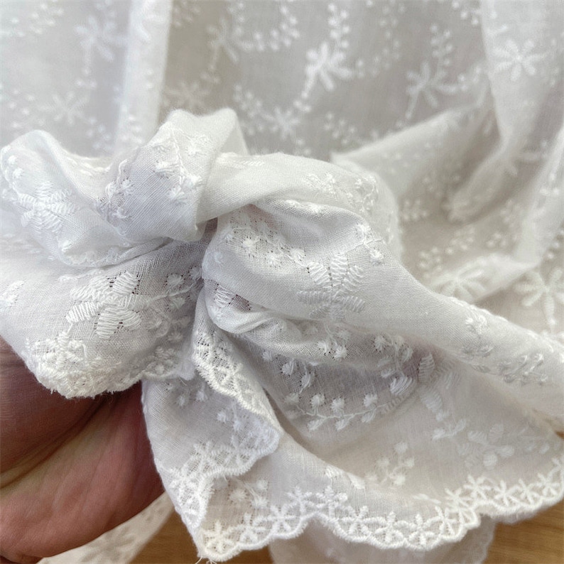 Puede incluir: Una tela blanca transparente con un intrincado patr&oacute;n floral bordado en toda la superficie. El material tiene un borde festoneado con un delicado acabado similar al encaje. La tela es ligera y semitransparente, mostrando un dise&ntilde;o bot&aacute;nico repetitivo.