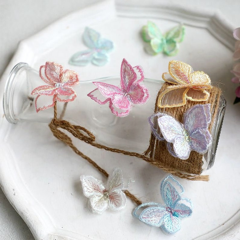 May include: Assortment of colorful embroidered butterfly appliques in various pastel shades, including pink, blue, yellow, and white. The appliques are arranged around a glass bottle with a brown twine wrap.